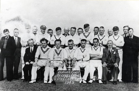 Photograph of twelve men in cricket whites accompanied by twelve other men in suits; the group is on grass, but nothing can be seen behind the men; in front of them is a notice reading as follows: Durham Coast Cricket League Div. 1 Champions 1949; the team has been identified as C. W. Cricket Club, South Hetton