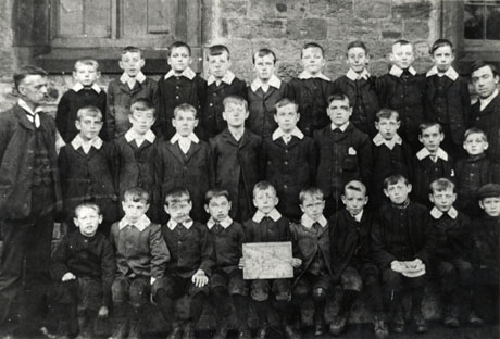 Photograph of a group of twenty eight boys, aged approximately eight years, standing against the wall of a stone building; the group is accompanied by two men, presumably schoolmasters, who have been identified as follows: Left: Mr. G. Lindsay; Right: Mr. T. Sexton; the school has been identified as the Colliery School at South Hetton