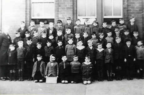 Photograph of a group of forty seven boys, aged approximately seven years, posed outside a building with long windows; on the left of the picture is a middle-aged man in a frock coat, presumably their teacher, identified as Mr. Lindsay; the group has been identified as being in South Hetton