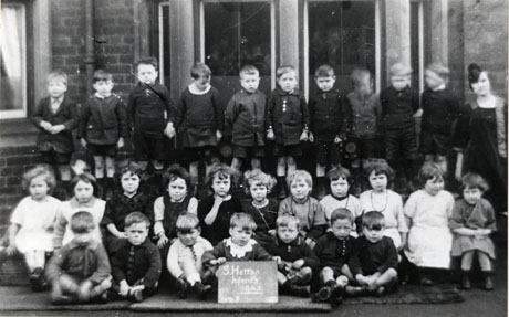 Photograph of twenty seven children, aged approximately six years, posed in front of a building with long windows; they are accompanied by a woman, identified as Miss Jones; the children have been identified as the infants class at South Hetton; a child on the front row is holding a slate bearing the words: S. Hetton Infants 1925