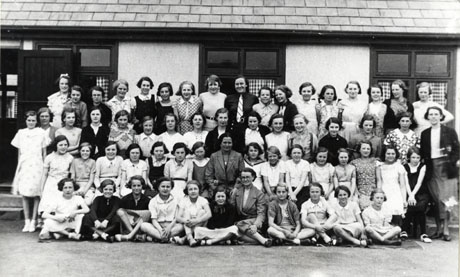 Photograph of a group of fifty three girls, aged approximately between thirteen and sixteen years, posed against the wall of a low building, possibly an accommodation block; they are accompanied by four adult women, presumably teachers; the girls are dressed in ordinary dresses, skirts, and shorts and have been identified as taking part in a school camp for pupils from South Hetton and Murton at Marske in Yorkshire