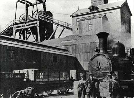 Photograph showing the winding gear and winding house of South Hetton Colliery with a further long low building at right angles to the winding house; standing beside the low building are a coal wagon bearing the markings SH 248 and a truck with the markings N E ; on the right of the picture is the front of a steam locomotive with three men standing in front of it