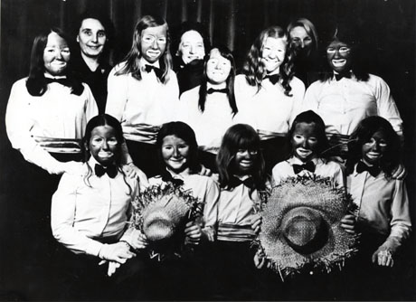 Photograph of ten girls, aged approximately fifteen years, wearing blouses, cummerbunds and bow ties and with their faces made up as Black and White Minstrels; two of the girls are holding straw hats; three women are standing behind the group, which has been identified as South Hetton Independent Methodist Chapel Minstrel Group