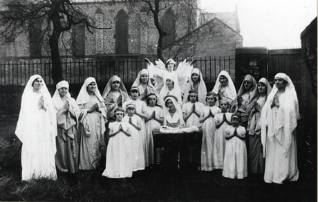 Photograph of eleven women and eight girls, aged approximately ten years, posed with the exterior of the church of Holy Trinity, South Hetton, behind them in the distance; the women and girls are dressed for their parts in a Nativity play; ten of the women, dressed in long robes, are standing in a semi-circle round the girls dressed as angels and another woman dressed as the Virgin Mary; behind the Virgin is another woman dressed as the Angel Gabriel; the Nativity play has been identified as being put on by South Hetton Mothers' Union