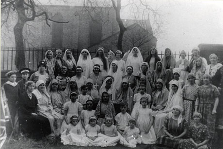Photograph of thirty five women, ten girls, and two boys posed in front of the exterior of the church of Holy Trinity, South Hetton, in the distance, in the costume of a Nativity play; the women are wearing long robes and scarves on their heads, apart from seven who are wearing conventional dress; the girls are dressed in angels' costumes and the boys are wearing military uniforms; the Virgin Mary is in the centre of the group; the play has been identified as being put on by the Womens' Institute in South Hetton
