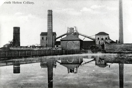 Postcard photograph entitled South Hetton Colliery showing three chimneys, three buildings and part of the winding gear reflected in the water of the reservoir near the colliery