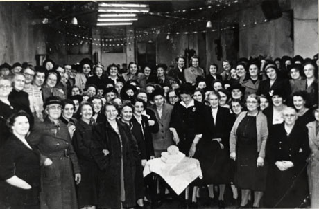 Photograph of approximately eighty women dressed in coats and hats standing in a hall surrounding a woman standing about to cut a large iced cake on a small table in front of her; they have been identified as members of South Hetton Women's Institute