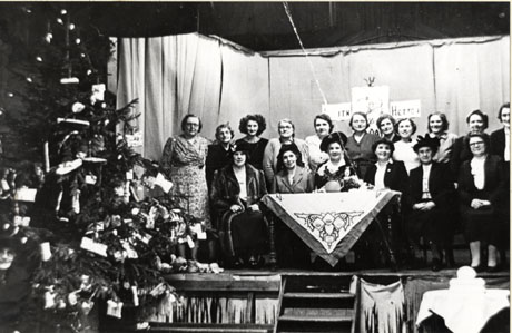 Photograph of thirteen women standing, and five women sitting, on a stage, with a table, on which there is a cloth, in front of them; to the left of the picture, is a large decorated Christmas tree in front of the stage; the group has been identified as the Committee of South Hetton Women's Institute, possibly presiding at the Institute's Christmas Party