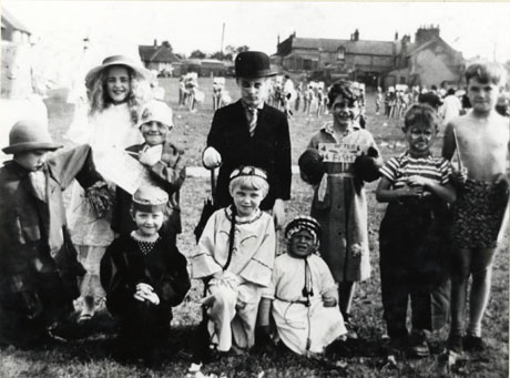 Photograph of a group of ten children, aged between approximately five and nine years, standing in a field in fancy dress with indistinct figures behind them; in the far distance a group of houses can be seen; one boy is dressed as a conventional business man in Bowler hat and with umbrella, but the costume of the other children is somewhat obscure; the occasion has been identified as South Hetton Welfare Carnival Day