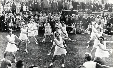 Photograph showing fourteen women in three lines facing the camera wearing short tunics, socks and tennis shoes, swinging their arms to the side; behind them are crowds of people watching them; the ground on which the women are standing appears to marked as a running track; the photograph has been described as Welfare Ladies' Group, South Hetton