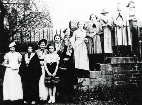 Photograph of twelve women and girls in costume standing on a flight of four steps in the open air with railings and an indistinct building behind them; the women are described as being members of an elocution class at South Hetton and may be dressed for a final show