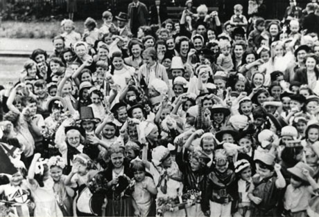 Photograph of a crowd of children and women photographed from above, showing that the majority are in fancy dress, including a small girl in the left hand corner who is wearing a costume bearing the symbol of the Festival of Britain; a Welsh hat can also be seen; the photograph has been described as Festival of Britain Carnival, South Hetton