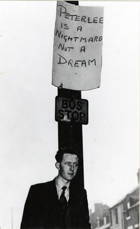 Photograph of an unidentified man standing in front of a bus stop on which a notice reading Peterlee is a Nightmare not a Dream; the photograph has been identified as recording part of the campaign by the inhabitants of South Hetton to remain outside the area of Peterlee, the New Town designated in 1948 and presumably with plans for expansion in 1959