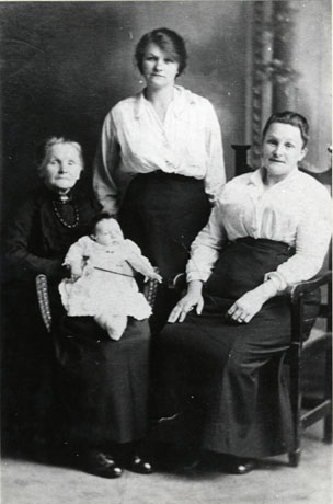 Photograph of an elderly woman sitting in an armchair at the left of the picture, holding an infant on her lap; she is dressed in a long dark dress and highly polished boots; in the middle of the photograph a young woman dressed in a light coloured blouse and a long dark skirt is standing; on the right of the picture a middle-aged woman also dressed in a light coloured blouse and a long dark skirt is sitting in a chair with arms; the group has been described as Four Generations of the Holmes Family of South Hetton