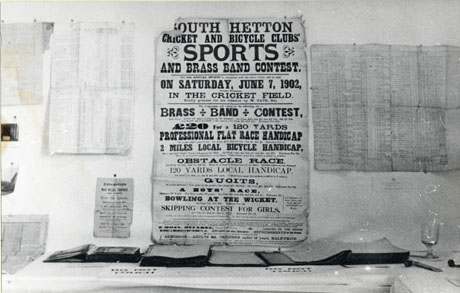 Photograph showing documents on display in an exhibition presumably concerning the history of South Hetton; six printed posters can be seen pinned on the wall; in front if the wall three open volumes can be seen lying on a table; only one of the printed posters can be read: a notice advertising South Hetton Cricket and Bicycle Clubs' Sports and Brass Band Contest on Saturday, June 7, 1902, in the Cricket Field; details of the programme for the day are given on the poster