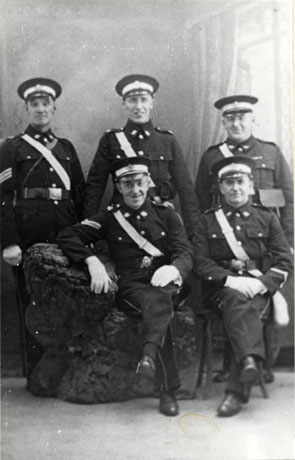 Photograph of three men standing and two men sitting, in the uniform of the St. John Ambulance Brigade, posed in what appears to be to be a photographer's studio; one man standing on the left has sergeant's stripes and one man sitting on the left has corporal's stripes; they have been described as being from South Hetton