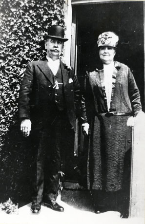 Photograph of a middle-aged man and woman standing against the doorway and wall of a house; the man, on the left, is wearing a bowler, a high collar, waistcoat, jacket, watch chain and striped trousers; the woman is wearing a toque hat, a drop-waisted dress of shiny material with a wavy pattern; they have been identified as Mr. Coscamp, Co-operative Secretary, and his Wife of South Hetton