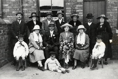 Photograph showing a group of five men and six women and three children posed in the yard of a terraced house against the wall of the house and the wall dividing the yard from that adjoining it; three women and four men are standing in a row behind one man, three women and three children, who are seated in front of them; one of the seated women is wearing a suit and holding a bouquet of flowers; the other women are dressed in suits; the men are also dressed in suits and hats; the small boys, aged approximately three to five years, are dressed in sailor suits; the event has been identified as Emma Barry's Wedding, South Hetton