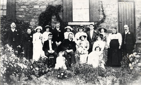 Photograph of six men, nine women and two small girls, posed against the wall of a stone house with plants in the garden of the house in front of them; the women are dressed in long skirts and large hats and the men in suits, but no hats; the girls, aged approximately five and six years, are dressed in light coloured dresses; the event has been identified as the wedding of Dick and Frances Holmes of South Hetton