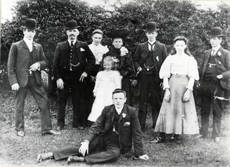 Photograph of four men wearing suits, ties and bowler hats standing with an elderly woman dressed in a large hat and dark frock, a middle-aged woman dressed in a light blouse and no hat, a girl aged approximately fifteen years in blouse and long skirt and girl aged approximately ten years in a light frock; in front of the group is a boy aged approximately sixteen years, wearing a suit and no hat, sitting on the ground; behind the group are bushes and there is grass under their feet; the group has been identified as The Holme(sic) Family of South Hetton