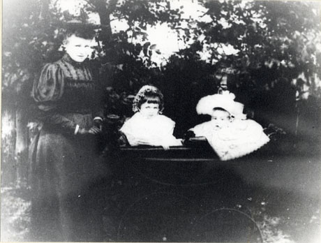 Photograph of woman wearing a high-necked frock with mutton sleeves pushing a perambulator containing an infant and a small girl, aged approximately two years; behind the them are trees; they have been identified as Miss Jenny Holmes with Dr. Kathleen and Dr. Donald Henega, of South Hetton, as children