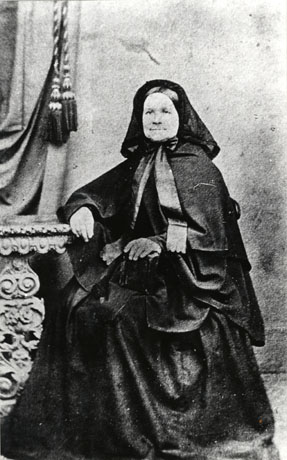 Photograph of a woman dressed in a voluminous crinoline-like skirt; an equally voluminous cape and a snood, on her head; she is sitting with her right arm resting on an elaborate table in a photographer's studio; it is possible that she is in mourning; she has been identified as Mrs. Tremelyn of South Hetton