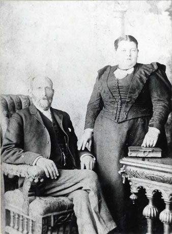 Photograph of a man seated on the left of the picture in an armchair, dressed in tie, jacket, waistcoat, watch chain and light coloured trousers; on the right a stout woman, wearing a dress of dark self-patterned fabric with small buttons down the front of the bodice, tight sleeves and two frills either side of the bust, is standing resting her right hand on the man's arm and her left on a book lying on a table; they have been identified as Mr. and Mrs. Tremelyn of South Hetton