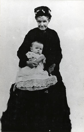 Photograph of and elderly woman standing in a long dark dress and dark hat holding an infant in elaborate lace-trimmed frock; they have been identified as Mrs Tremelyn and grandchild, South Hetton