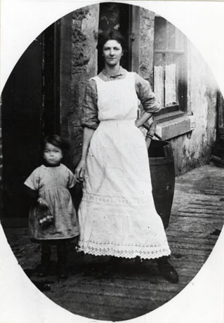 Photograph of a young woman dressed in a long light-coloured pinafore to her ankles, holding the hand of a small girl, aged approximately five years, dressed in a pinafore to her knees, and carrying a little bag; behind the pair are the wall, window and doorway of a roughly built stone house; part of a poss tub can be seen behind the woman; under their feet is rough concrete; they have been identified as Mrs. Tremelyn and daughter, Smith Street, South Hetton