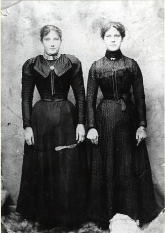 Photograph of two young women standing side by side facing the camera; they are both dressed in dark frocks with long sleeves, narrow corseted waists and chains round their necks; they have been identified as: Left; Mrs. Shipley; Right: Mrs. Rodgers of South Hetton