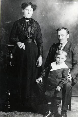 Photograph showing a woman standing on the left of the picture wearing a long dark frock with long sleeves, a narrow waist and a chain round her neck, leaning on a wooden stand; next to her is a man wearing a suit and tie sitting on a chair with a small boy, aged approximately six years, standing at his knee; the boy is dressed in a suit with short trousers and a large collar; they have been identified as the Rodgers Family of South Hetton