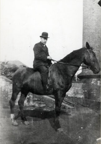 Photograph of a man sitting on a horse which is standing with its side to the camera; the man is wearing a suit and bowler hat; behind him, on the left of the picture, is what seems to be part of a large chimney; he has been identified as Mr. Holbourn, Head Horse Keeper, South hetton Colliery