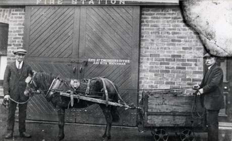 Photograph showing a pony, wearing blinkers, harnessed to a coal waggon in front of the door and wall of a fire station; a man dressed in a suit and cap and identified as Mr. Coates, is standing on the left of the picture, holding the pony by a rein; a man wearing a suit and cap is standing on the right of the picture holding the pony's reins across the coal waggon; he has been identified as Mr. Holbourn, Head Horse Keeper at South Hetton Colliery