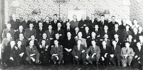 Photograph of fifty one men posed against a wall covered in wallpaper on which a large mirror is hanging; the men who are all wearing suits are described as South Hetton Colliery Officials; the man in the centre of the group has been identified as Mr. Mackie, Manager