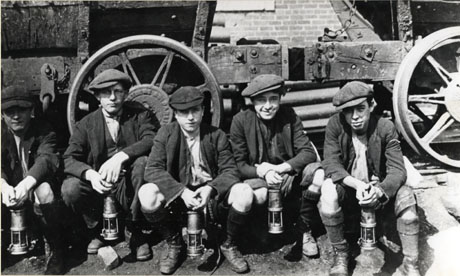 Photograph of five young boys wearing caps, jackets, long socks and boots and carrying miners' lamps, squatting in front of a coal waggon; they have been identified as miners at South Hetton, and specifically as follows: Second Left: Norman Bell; Far Right: Mr. Macdermott