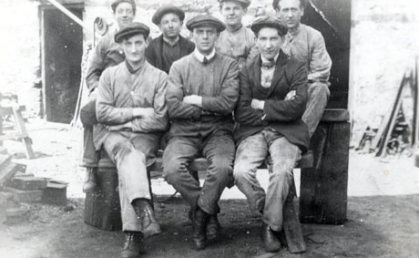 Photograph of three young men sitting on a bench with four young men standing behind them; behind the group a wall in the distance; pieces of wood and bricks lying about can be seen; the men are wearing overalls and caps and have been identified as miners at South Hetton Colliery