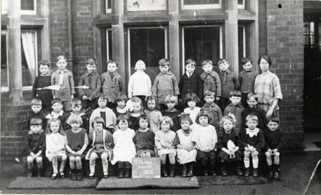 Photograph of nineteen boys and fifteen girls, aged approximately six years, grouped in front of a bay window in a large building; a woman, presumably their teacher, is standing at the right of the picture; a notice at the front of the picture reads: South Hetton Infants, 1925