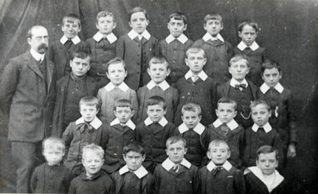 Photograph of twenty four boys, aged approximately nine years, grouped in front of fabric; a man with a walrus moustache, identified as Mr. Blamire, is standing at the left of the picture; the children have been identified as pupils at South Hetton