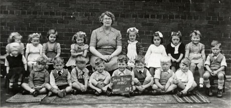 Photograph of nine girls and nine boys, aged approximately four years, sitting in two rows, with a middle aged woman sitting in the middle of them; the woman, who has been identified as Miss Alcroft, is wearing a dress with short sleeves; all the children are dressed in summer clothing; a child on the front row is holding a notice reading: South Hetton Infants Babies 1949