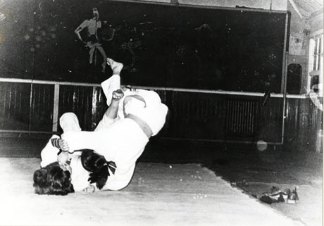 Photograph of two people in white martial arts garments falling locked together on a mat; the faces of neither individual can be seen; behind the mat a screen with a stylised picture of an oriental warrior can be seen; the picture has been identified as The Judo Club in the Youth Club, South Hetton