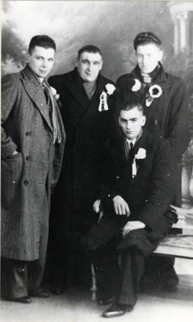 Photograph of three young men in overcoats standing behind a fourth young man sitting on a bench in a photographer's studio; all the men are wearing rosettes and button holes; they have been identified as South Hetton Fans Off To Sunderland Cup Final, 1937