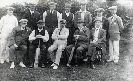 Photograph of twelve men posed in front of bushes; five of the men are wearing cricket whites; the man in the middle of the front row is wearing a striped cap; two men on the front row are holding cricket bats; they have been identified as South Hetton Cricket Team, circa 1910