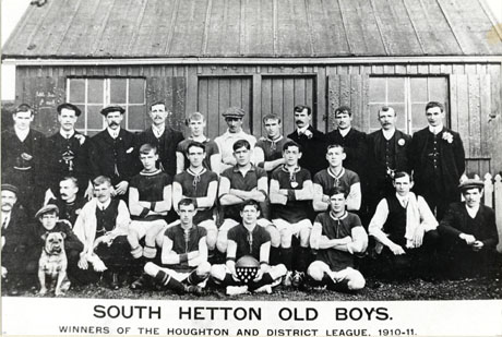 Photograph of eleven men in football strip, accompanied by fourteen other men and a dog, posed in front of a low wooden building; a player on the front row is holding a football and a shield containing eleven medals; the picture is captioned as follows: South Hetton Old Boys, Winners of The Houghton and District League, 1910-11