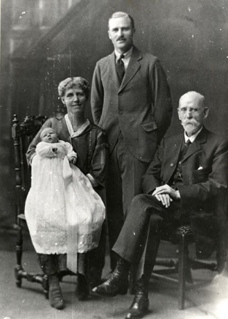 Photograph of a young man wearing a suit and tie, standing between a woman sitting on a chair, holding a baby in a long elaborate robe, possibly a christening robe; on his other side, an elderly man, wearing a suit and tie, is sitting on a chair; behind the group, what appears to be the balustrade of a staircase can be seen; the photograph has been identified as W. O. Wood (right) Manager of South Hetton Colliery, with his family