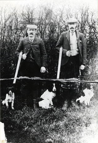 Photograph of two men standing facing the camera with shotguns broken over their arms; three dogs, two spaniels and a terrier, are sitting at their feet; both men are wearing jackets, ties, waistcoats, caps and moustaches; behind them leafless trees can be seen; they have been identified as Mr. Stubbs and a local gamekeeper in South Hetton