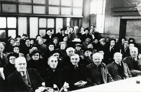 Photograph of approximately fifty elderly people wearing overcoats sitting in rows in a hall; a man at the back of the group is holding what appears to be a large iced cake; the photograph has been identified as Senior Citizens Party, South Hetton