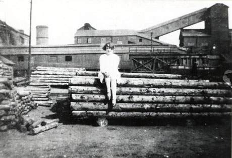 Photograph of a young woman sitting on a pile of logs, possibly pit props, looking towards the camera, behind her are further piles of logs and the buildings of the colliery identified as South Hetton Colliery