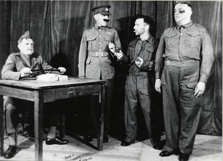 Photograph of the cast of a production of Stars in Khaki, showing a man in army uniform sitting at a table at the left of the picture; a man in army uniform with a moustache and baton, possibly a sergeant major, is standing next to the table; next to him is a smaller man in uniform, possibly a private soldier; at the right, is a fourth man in army uniform with a black eye; the cast are on a stage