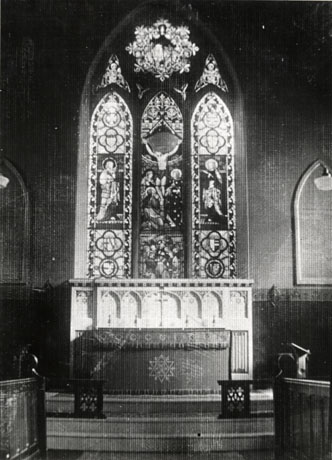 Photograph of the interior of a church showing the East window with stained glass, depicting the crucifixion, unidentified saints and coats of arms; the altar and altar steps can also be seen; the church has been identified as the church of Holy Trinity, South Hetton