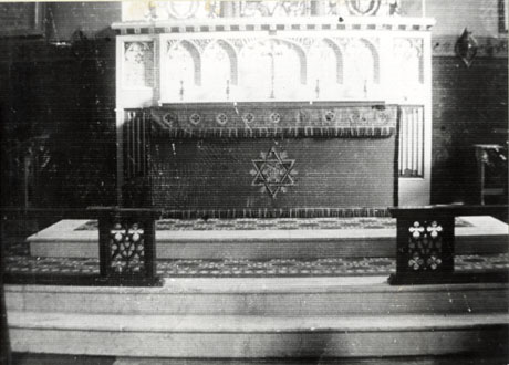 Photograph showing the altar frontal, altar steps, and reredos in a church, which has been identified as Holy Trinity, South Hetton; the frontal has a Star of David and IHS on it; the reredos consists of arches carved in stone
